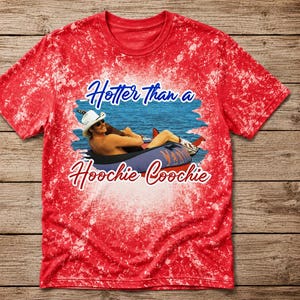 Könnte beinhalten: Ein rotes T-Shirt mit Bleicheffekt und einem Grafikdruck einer Person mit weißem Cowboyhut und Sonnenbrille, die in einem blauen Reifen auf dem Wasser treibt. Der Text Hotter than a Hoochie Coochie steht in blauer und roter Schrift darauf.