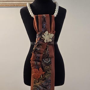May include: A necktie accessory featuring a brown and black paisley patterned fabric with solid brown ribbed sections. The piece is adorned with two leaf-shaped brooches, one gold and one orange, and is suspended by a white pearl necklace.