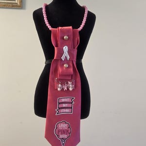 May include: A pink fabric necktie featuring a pearl bead necklace, a silver ribbon pin, and two embroidered patches. The top patch reads "SWEET BUT STRONG" and the bottom patch is shaped like an ice cream cone with the text "LOOK ON THE PINK SIDE."