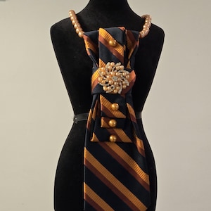 May include: A necktie accessory featuring a navy blue, brown, and gold striped pattern. The tie is adorned with a central circular brooch made of clear crystals and pearls, accompanied by several round, metallic gold beads.