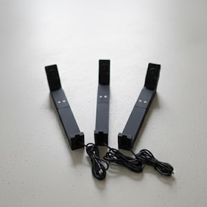 Könnte beinhalten: Drei schwarze Ständer für elektronische Geräte mit angebrachten schwarzen Kabeln. Jeder Ständer hat eine rechteckige Basis und einen vertikalen Arm mit einer kleinen, abgewinkelten Oberseite. Die Ständer sind auf einer hellgrauen Oberfläche angeordnet.