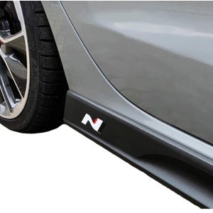 以下が含まれることがあります： シルバーの車に取り付けられた黒いサイドスカート。スカートには白と赤の「N」ロゴが付いています。車のタイヤとホイールが背景に見えます。