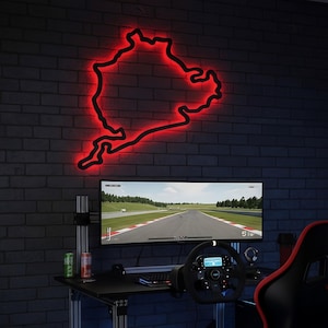 Puede incluir: Un contorno rojo neón de un mapa de circuito de carreras está montado en una pared de ladrillos oscuros. Debajo, una configuración de juego incluye un monitor grande que muestra un juego de carreras, un volante y un escritorio negro.