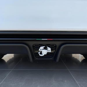 Könnte beinhalten: Nahaufnahme des hinteren Stoßfängers eines Autos mit einem schwarzen Wabenmuster-Grill und einem weißen Skorpion-Emblem. Der Stoßfänger hat eine schwarze Zierleiste mit einem kleinen italienischen Flaggen-Detail. Die Karosserie des Autos ist weiß.