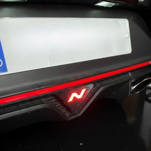 Könnte beinhalten: Nahaufnahme des hinteren Stoßfängers eines Autos mit einem leuchtenden roten "N"-Logo. Der Stoßfänger ist schwarz mit einem roten Zierstreifen. Ein weißes Nummernschild ist über dem Logo zu sehen.