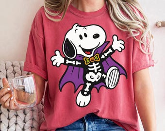 May include: A red short-sleeve t-shirt featuring a graphic of a white cartoon dog dressed as a skeleton with a purple cape and an orange bow tie with a pumpkin face. The dog has its arms outstretched and is smiling against the red fabric background.