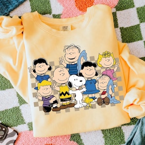 May include: A pale yellow crewneck sweatshirt featuring a graphic print of the Peanuts characters including Charlie Brown, Snoopy, Woodstock, Lucy, Linus, Sally, and Schroeder over a grey and white chequered background with a Comfort Colors label.