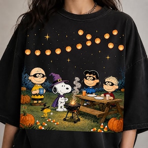 May include: A black t-shirt featuring a graphic of Peanuts characters celebrating outdoors at night. The scene includes Snoopy in a purple witch hat, Charlie Brown, Lucy, and Linus near a campfire, pumpkins, and string lights against a dark sky.