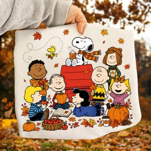 May include: A white sweatshirt featuring a colorful Peanuts graphic with Snoopy, Charlie Brown, and friends surrounded by autumn leaves, pumpkins, and a basket of apples. The characters are gathered around a red doghouse in a fall setting.