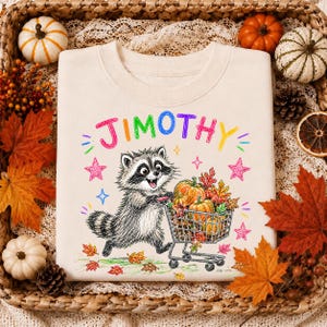 Puede incluir: Una sudadera de cuello redondo color crema con un gráfico de un mapache empujando un carrito de compras lleno de calabazas y hojas de otoño. El nombre JIMOTHY está impreso en letras coloridas estilo crayón sobre el mapache, rodeado de estrellas.