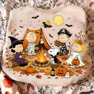 以下が含まれることがあります： ベージュのクルーネックトレーナーに、ピーナッツのカラフルなハロウィンのグラフィックが描かれています。テント、カボチャ、コウモリ、満月、秋の葉が散りばめられたキャンプファイヤーの周りで、仮装したキャラクターたちが描かれたデザインです。