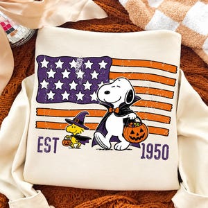 May include: A cream-colored sweatshirt featuring a graphic of Snoopy dressed as a vampire and Woodstock as a witch, both holding jack-o'-lantern buckets. Behind them is a distressed American flag in purple and orange. The text reads EST 1950.