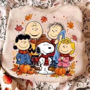 May include: A beige crewneck sweatshirt featuring a colourful graphic of the Peanuts characters Charlie Brown, Lucy, Linus, Sally, and Snoopy wearing scarves and hats, surrounded by orange and red autumn leaves and two orange pumpkins.