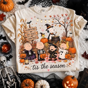 Op de afbeelding: Een crèmekleurig t-shirt met een afbeelding van Peanuts-personages in Halloween-kostuums. Het ontwerp bevat een houten bord met de tekst PUMPKINS, HAY-RIDES, CORN MAZE, een lichtsnoer en de tekst 'tis the season aan de onderkant.