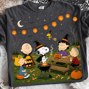 May include: A charcoal gray t-shirt featuring a Peanuts Halloween graphic. The design shows Charlie Brown, Snoopy, Woodstock, Lucy, and Linus in costumes at night under string lights, a crescent moon, and stars, with pumpkins and a grill.