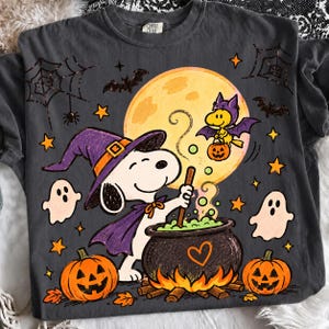 May include: A charcoal gray t-shirt featuring a graphic of a white dog wearing a purple witch hat and cape, stirring a black cauldron with green bubbles. A yellow bird dressed as a bat flies above near a large moon, surrounded by ghosts and pumpkins.