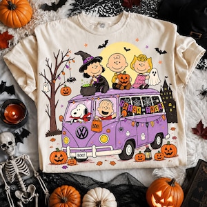 May include: A cream-colored t-shirt featuring a graphic of Peanuts characters in a purple van decorated for Halloween. The design includes Snoopy, Charlie Brown, Lucy, and Linus with pumpkins, bats, and a banner that reads TRICK OR TREAT.