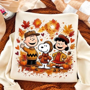 May include: A cream-colored sweatshirt featuring a graphic print of Charlie Brown, Snoopy, and Lucy wearing autumn clothing. The characters are surrounded by colorful fall leaves, small pumpkins, acorns, and hearts in shades of orange, red, and brown.