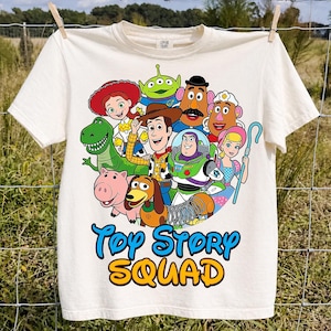 Può includere: Una t-shirt color crema con una grafica colorata dei personaggi di Toy Story, tra cui Woody, Buzz Lightyear, Jessie, Rex, Slinky Dog, Hamm, Mr. Potato Head, Mrs. Potato Head, Bo Peep e un Alien. Il testo "Toy Story SQUAD" è stampato sotto.