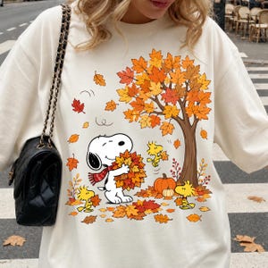 Op de afbeelding: Een crèmekleurig sweatshirt met ronde hals en een grafische print van Snoopy met een rode sjaal die een hoop herfstbladeren vasthoudt. Het ontwerp bevat een grote boom met oranje en geel gebladerte, twee gele vogels en pompoenen.
