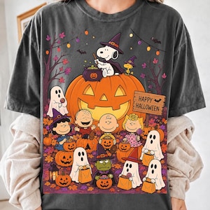 May include: A charcoal gray t-shirt featuring a graphic print of Peanuts characters in Halloween costumes. Snoopy sits on a large jack-o'-lantern, while Charlie Brown, Lucy, and others collect candy. A wooden sign reads Happy Halloween.