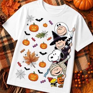 May include: A white short-sleeve t-shirt featuring a Halloween-themed graphic print. The design includes orange jack-o'-lanterns, black bats, spiderwebs, autumn leaves, and colorful candies alongside Peanuts characters dressed in costumes.