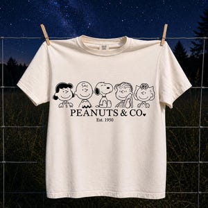 May include: A cream-coloured short-sleeve t-shirt featuring a black line-art graphic of five Peanuts characters. Below the characters, the text reads PEANUTS & CO. Est. 1950. The shirt has a crew neckline and is displayed hanging from two wooden clothespins.