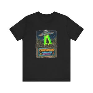 Puede incluir: Camiseta negra con un gráfico de un OVNI que emite un rayo verde sobre un árbol. El diseño incluye el texto "LEAVE NO TRACE CAMPGROUND STARGAZER RETREAT". La imagen tiene una estética vintage y de exteriores.
