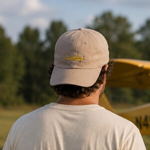 Könnte beinhalten: Eine hellbeige Baseballkappe mit einer kleinen, gestickten Grafik eines gelben Propellerflugzeugs in der vorderen Mitte. Die Kappe hat eine strukturierte Krone, einen gebogenen Schirm und einen Knopf oben, gefertigt aus einfarbigem Stoff.
