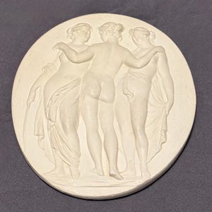 May include: A circular, off-white relief sculpture showing three figures in classical dress. The figures are intertwined, with two figures embracing the central figure. The sculpture is set against a dark grey background.