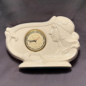 May include: Cream-colored decorative clock with a gold-tone clock face. The clock features a relief of a woman's profile and ornate detailing. The clock is approximately 7 inches wide and 5 inches tall.