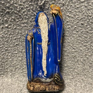 Peut inclure: Figurine d'un magicien en longue robe bleue avec une barbe blanche et un bâton. Le magicien lève un objet doré. La base de la figurine est marron et dorée.