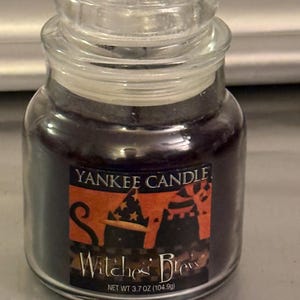 May include: A small Yankee Candle jar candle in the scent Witches' Brew, featuring dark purple wax and a clear glass lid. The label displays an orange and black graphic with a witch hat and the text Yankee Candle Witches' Brew Net Wt 3.7 oz (104.9g).