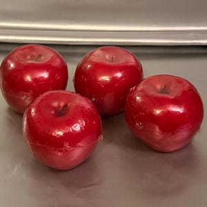 May include: Four glossy, bright red apple-shaped objects arranged in a cluster. Each piece features a smooth, reflective surface with a small indentation at the top, mimicking the appearance of a real fruit. The items are wrapped in a thin, clear plastic film.