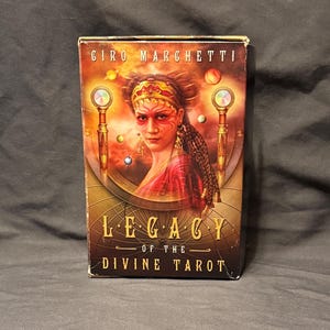 May include: A tarot card deck box featuring the title "Legacy of the Divine Tarot" and the artist's name, "Giro Marchetti." The box art depicts a woman with face paint and a red shawl, with gold and orange accents.