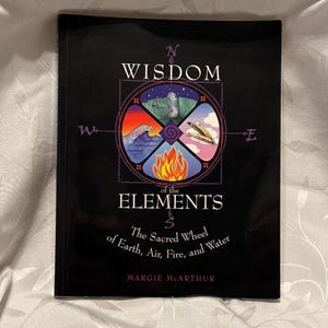 May include: A hardcover book titled "Wisdom of the Elements" by Margie McArthur. The cover is black with a central circular design featuring symbols representing earth, air, fire, and water. The book's title and author's name are in white.