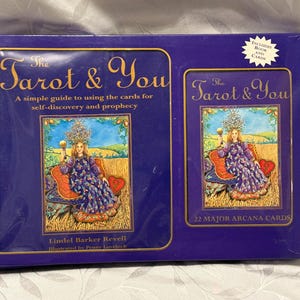 Op de afbeelding: Een doos met kaarten en een boek "The Tarot & You". De donkerblauwe doos heeft gouden randen en illustraties van een vrouw in een paarse jurk. De tekst luidt: "Een eenvoudige gids voor het gebruik van de kaarten voor zelfontdekking en profetie."