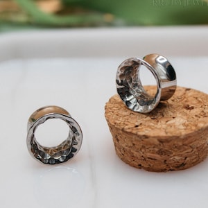 Op de afbeelding: Twee zilverkleurige oor stretchers met een gehamerde textuur worden getoond. Eén ligt op een kurk, de andere apart. De stretchers hebben een gepolijste afwerking. De achtergrond is wazig.