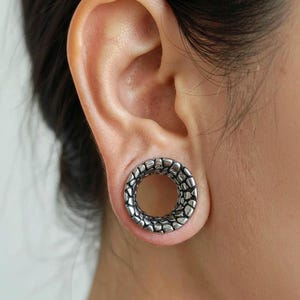 May include: A silver-colored ear gauge with a textured, cobblestone-like design. The gauge is circular and worn in a stretched earlobe. The jewelry is a tunnel style, allowing visibility through the ear.