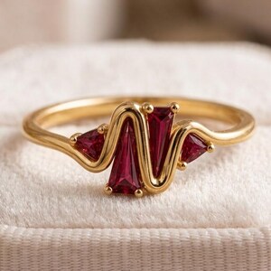 May include: A gold ring featuring a wavy, serpentine band set with four deep red, trapezoidal-cut gemstones. The metal band curves around each stone, creating a continuous, fluid design that highlights the vibrant crimson color of the gems.