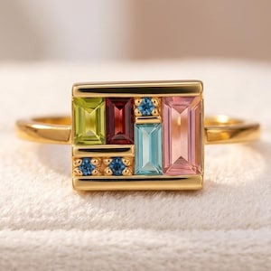 Pode incluir: Um anel de ouro com uma peça central retangular cravejada com uma variedade de pedras preciosas multicoloridas. O arranjo inclui pedras de corte baguette em verde-lima, vermelho-escuro, azul-claro e rosa-suave, acentuadas por quatro pequenas pedras azuis redondas.