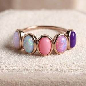 May include: A gold-toned ring featuring five oval-shaped cabochon stones arranged in a row. The stones include a pale lavender, a white opal with iridescent flashes, a solid pink, a pink opal with iridescent flashes, and a deep purple stone.