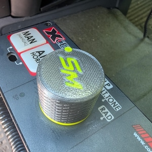 May include: A cylindrical silver knob with a textured surface and a bright neon yellow top featuring the letters SM. The knob is positioned on a black control panel with white text labels including MAN, MANUAL, AL, HORN, SP, P, LT.TONE, and RAD.