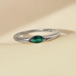 Peut inclure: Une bague en argent ornée d'une pierre précieuse verte émeraude de forme marquise. L'anneau poli présente une légère courbe. La pierre est sertie dans un sertissage à griffes. La bague est présentée sur un fond neutre.