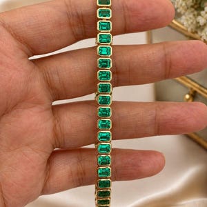 May include: A gold bracelet featuring emerald-cut green gemstones. The bracelet is set in a gold band and is displayed against a neutral background. The gemstones are rectangular and evenly spaced.