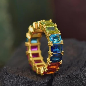 May include: A gold ring set with a rainbow of square-cut gemstones. The ring showcases a vibrant spectrum of colours, including red, orange, yellow, green, blue, and purple. The gemstones are set in a gold band, creating a striking and colourful piece of jewellery.