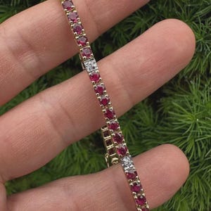 May include: A gold bracelet featuring a line of oval-cut, deep red gemstones. The bracelet includes a small section of white gemstones. The bracelet is set against a backdrop of green foliage.
