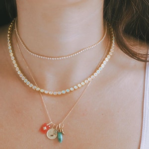 Puede incluir: Un conjunto de collares en capas con tres cadenas doradas. La cadena superior es una delicada hebra de pequeñas piedras transparentes. La cadena central tiene piedras de ópalo y la cadena inferior tiene colgantes.