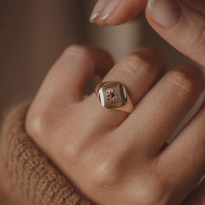 Könnte beinhalten: Ein goldener Siegelring mit quadratischer Oberfläche, der ein Sternmuster und einen kleinen roten Edelstein in der Mitte aufweist. Der Ring wird an einem Finger getragen, mit einem weichen, neutralen Hintergrund.