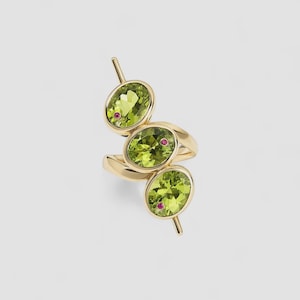 Puede incluir: Un anillo de oro con tres piedras preciosas ovaladas de color verde claro, cada una con un pequeño acento rojo. Barras doradas se extienden de cada piedra preciosa. El anillo se muestra sobre un fondo blanco liso.